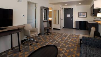 A hotel room with a TV, a desk, a chair, a sofa, a kitchen area, and a patterned floor.