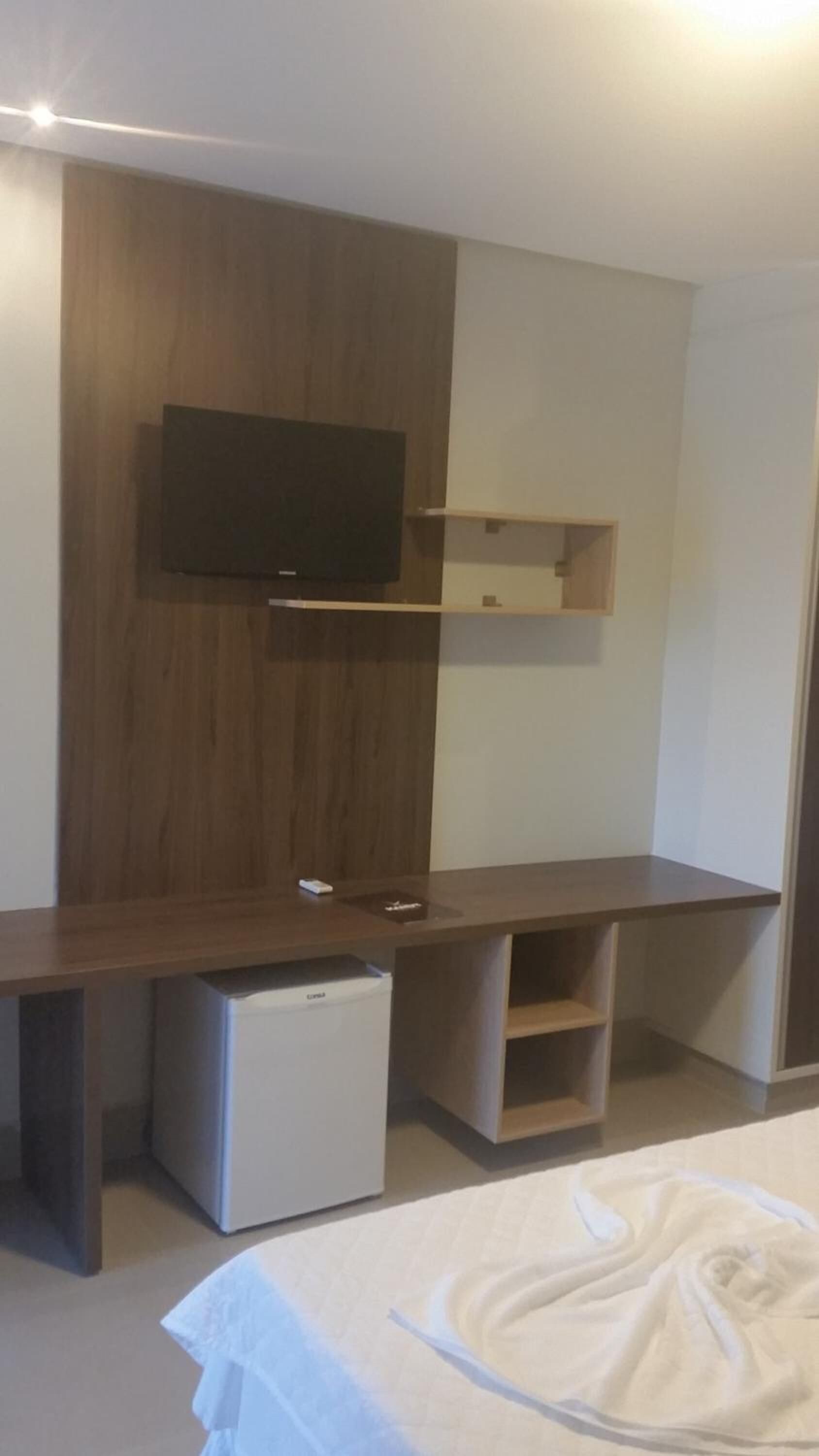 A modern room with a wooden desk, a flat-screen TV mounted on the wall, a small white refrigerator, and a bed with a white blanket.