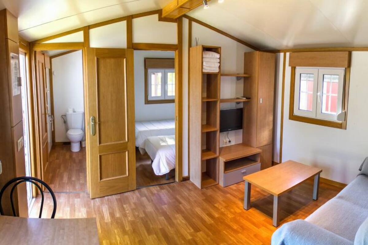 A compact living space with a wooden floor, a sofa, a TV, a small table, and a bathroom with a toilet and a bed.