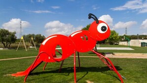 Una escultura de hormiga roja en un parque con césped, árboles y cielo despejado.
