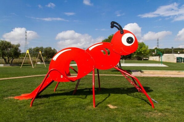 A red ant sculpture in a grassy park area with trees and a clear sky.