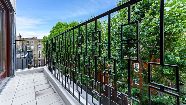 Varanda com grade de metal preta, piso de azulejos brancos e vista para um edifício de tijolos vermelhos e vegetação.