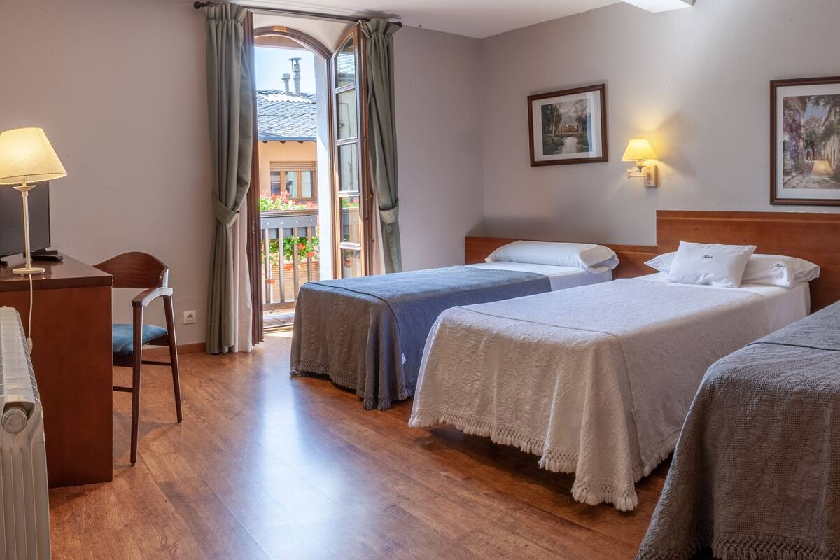 A hotel room with three beds, a desk with a lamp, and a view of the outside.