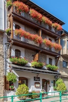 Un edificio tradicional europeo con varios balcones decorados con flores rojas, un letrero que indica un restaurante y una barandilla verde en primer plano.