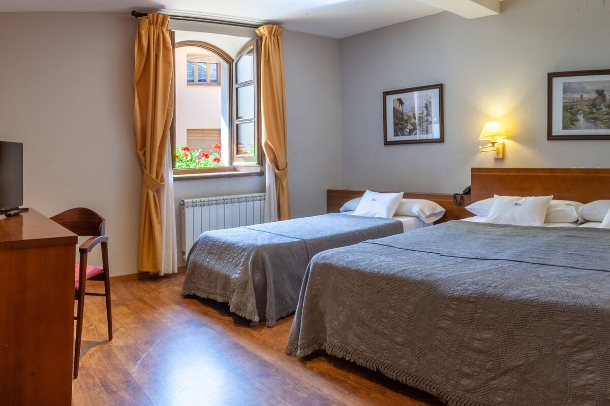 A hotel room with two beds, a wooden desk, a chair, and a window with curtains.