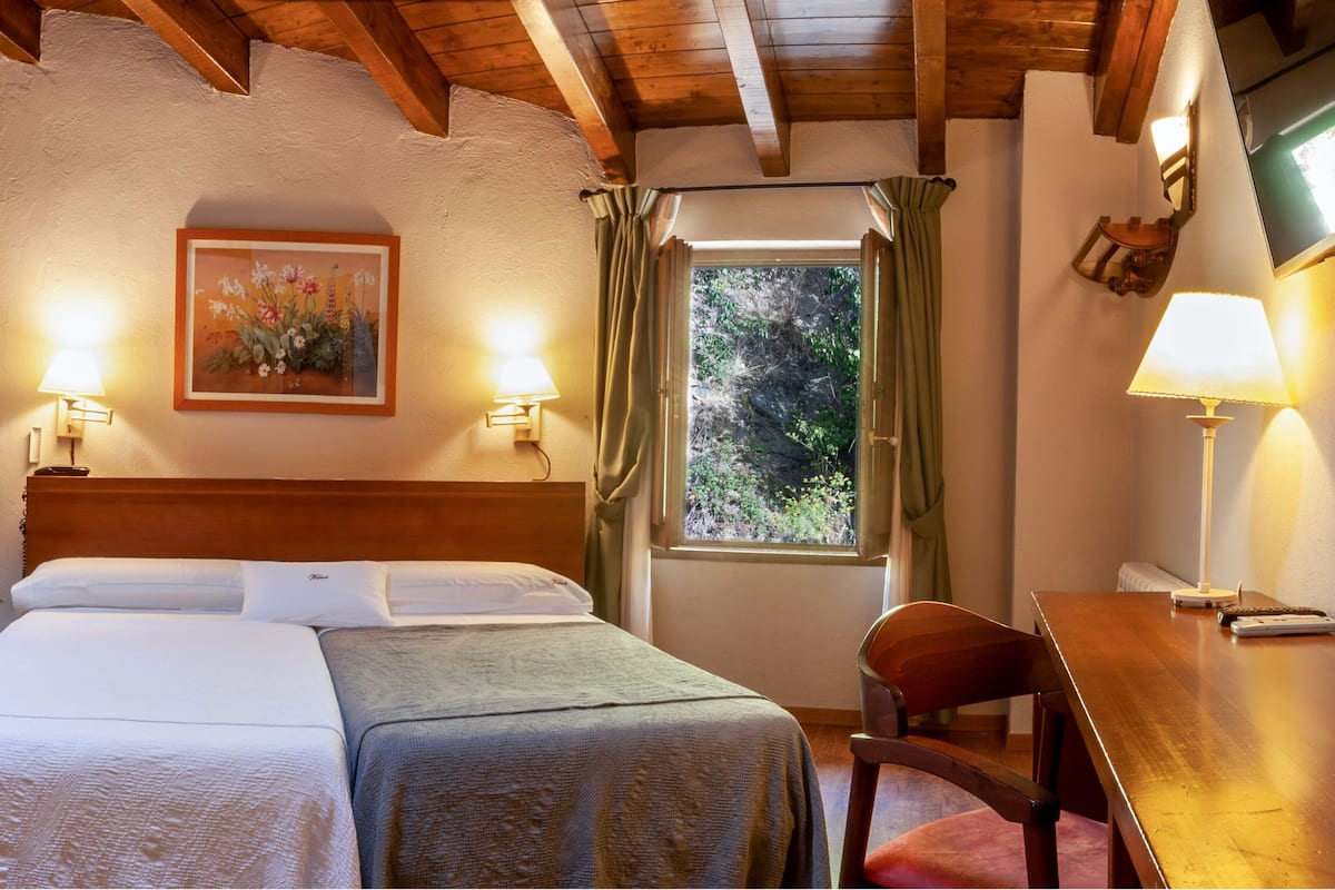 A room with a wooden staircase, a bed with two pillows, a bedside table with a lamp, and a window with curtains.