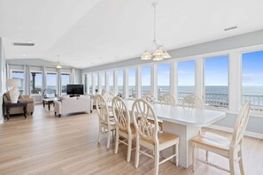 Un amplio espacio de sala con una gran mesa de comedor, múltiples sillas, un sofá, un televisor y un balcón con vistas al océano.