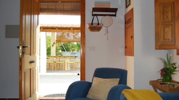 Una habitación con un sillón azul, una puerta de madera, un estante pequeño con una cesta y un estante en la pared con un atrapasueños.