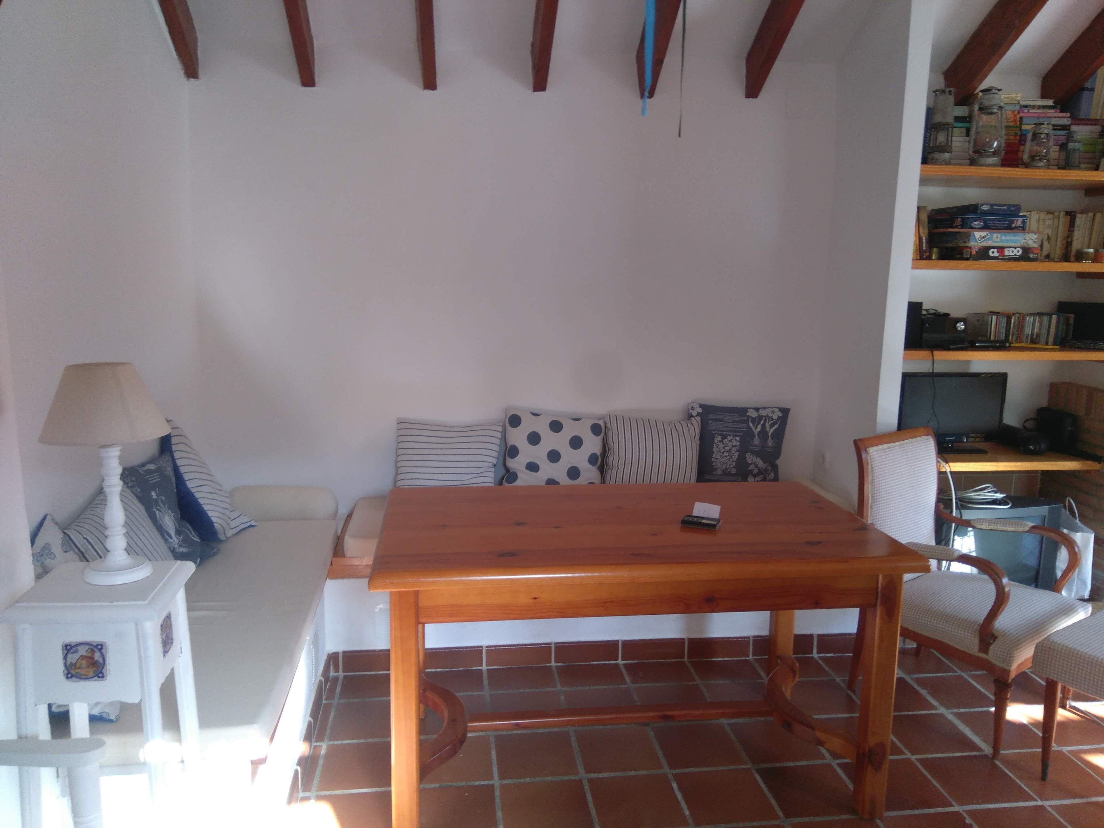 A living room with a wooden table, a sofa with patterned cushions, a small white side table with a lamp, and a bookshelf filled with books and decorative items.