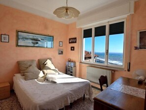 Un dormitorio con cama, escritorio, silla, radiador, ventana con vista al mar y cuadros enmarcados en la pared.