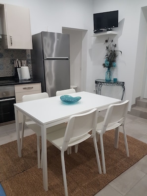 A compact kitchen with white cabinets, a stainless steel refrigerator, a white dining table with four chairs, and a small side table with a vase.