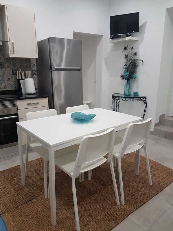 A compact kitchen with white cabinets, a stainless steel refrigerator, a white dining table with four chairs, and a small side table with a vase. - Casa Azul - Monte da Caldeirinha (Tavira)