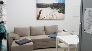 Een moderne woonkamer met een beige bank, een glazen tafel en een witte eettafel. Aan de muur hangt een schilderij van een landschap en boven de bank staat een airconditioning.