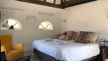 Un dormitorio con cama, dos ventanas, una silla y una mesita de noche.
