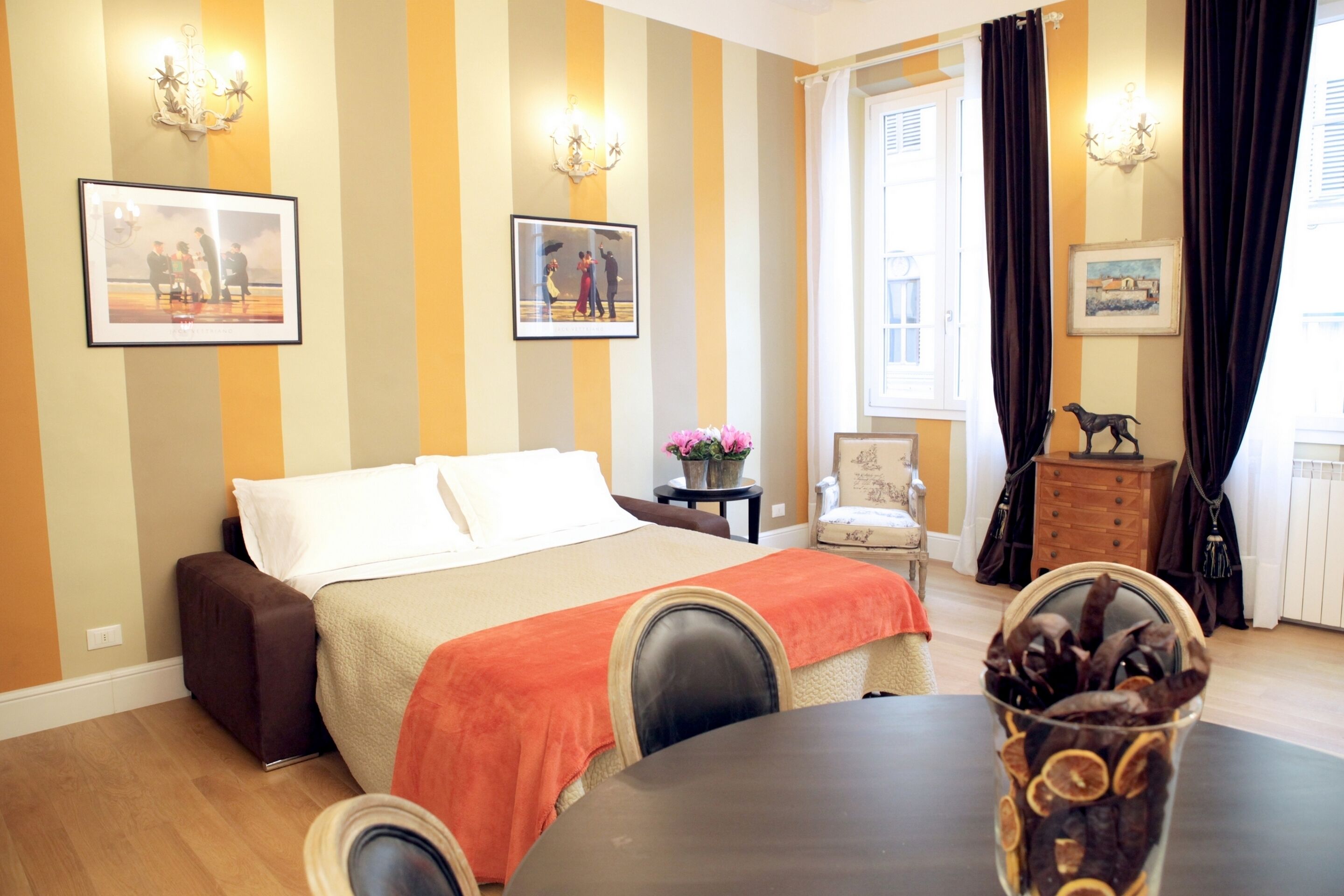 A hotel room with a bed, a table with a vase of dried flowers, a chair, and a dresser.