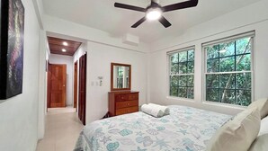 Une chambre à coucher comprenant un lit, un ventilateur de plafond, une commode, un miroir et une fenêtre donnant sur des arbres.