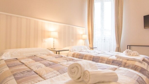 A hotel room with two beds, each with a quilted bedspread, bedside lamps, and rolled towels on the beds. - Appartamento Cittadella (Florence)