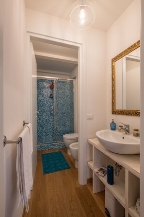 A bathroom with a white sink, a mirror with a gold frame, a blue shower with mosaic tiles, and a wooden floor. - Appartamento Parione (Florence)