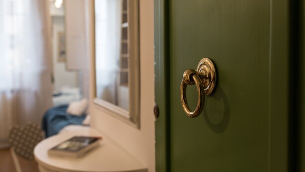 Une pièce avec une porte verte, une table ronde sur laquelle se trouve un livre, et un lit recouvert d’une couverture bleue.