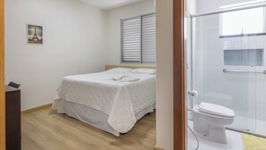 Un dormitorio con cama, mesita de noche, ventana con persianas y baño con inodoro y ducha.