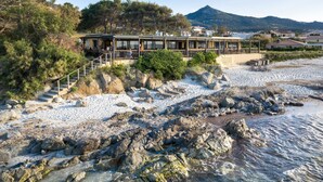 A coastal hotel with outdoor seating, a staircase leading to the beach, and a mountain in the background. - Joseph Charles (L'Île-Rousse)