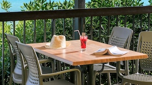 A wooden table with a hat and a drink, surrounded by chairs, overlooking a clear blue sea with a ship in the distance. - Joseph Charles (L'Île-Rousse)