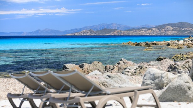 A beach with clear turquoise water, rocky shoreline, and mountains in the background. There are three sun loungers on the sand. - Joseph Charles (L'Île-Rousse)