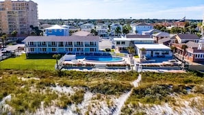 A residential area with a large house featuring a swimming pool, surrounded by other houses and a beach in the background.