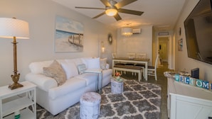 A living room with a white sofa, a coffee table, a lamp, a ceiling fan, a painting, a chandelier, a dining area, and a welcome sign.