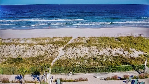 A beach with a clear view of the ocean, a sandy shore, and a grassy area with a fence and chairs.