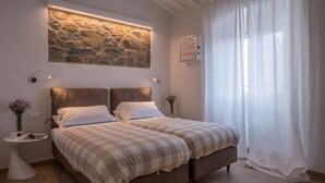 A double bed with checkered bedding, a stone wall with a light fixture, a bedside table with a vase, and a window with sheer curtains. - Appartamento Matteotti (Florence)