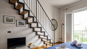 A modern bedroom with a blue bed, a TV on a white desk, a staircase with wooden steps, and framed pictures on the wall. - Apollonia Apartment (Florence)