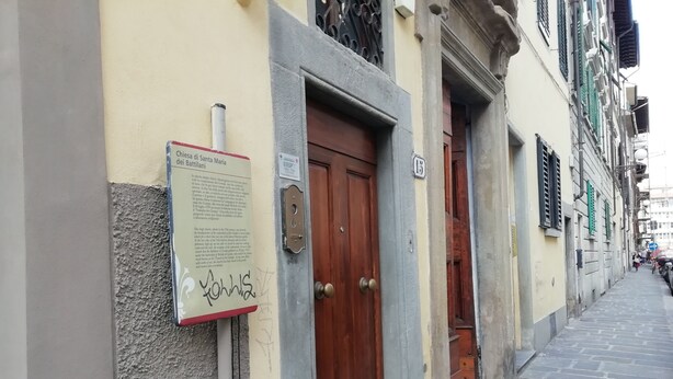 A narrow street with a building featuring a wooden door and a plaque with text. - Appartamento Ruote (Florence)