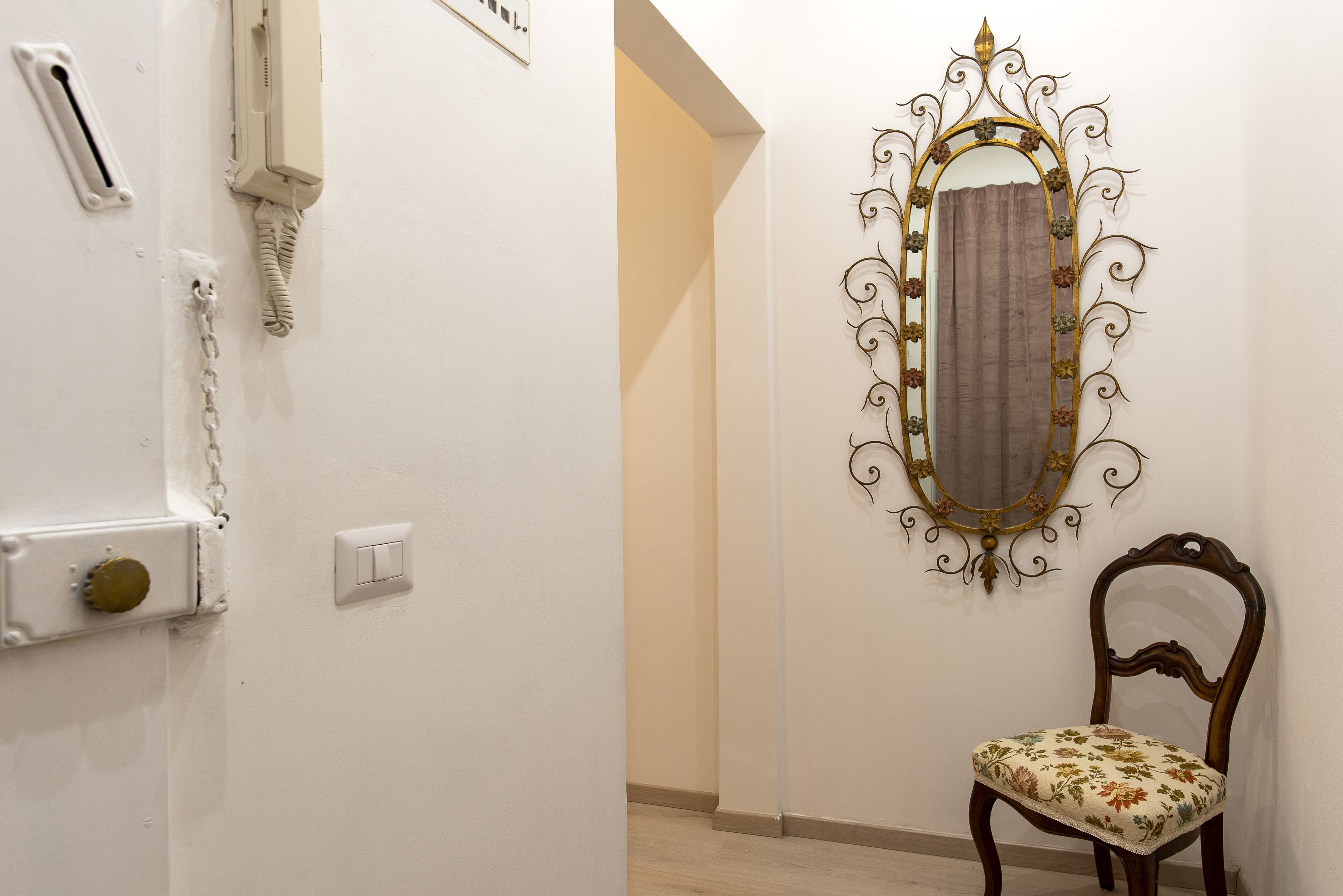Une pièce avec un mur blanc, un miroir décoratif, une chaise en bois et un interrupteur.