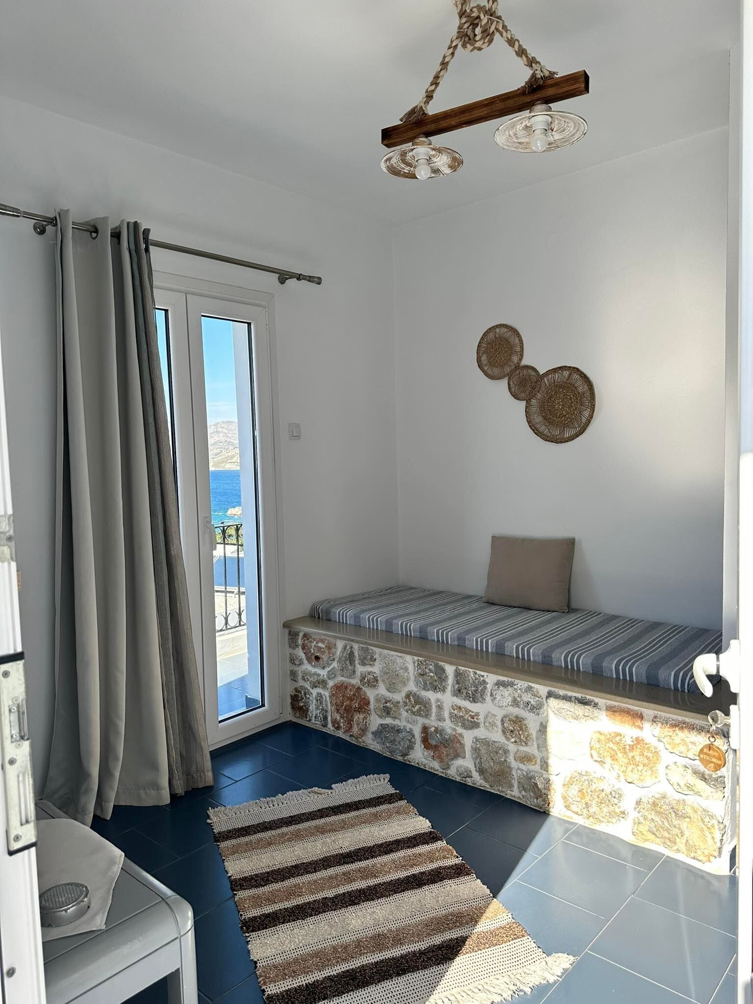 Una habitación con pared de piedra, cama con cubrecama a rayas, alfombra tejida, lámpara de techo y vista al mar a través de una ventana.