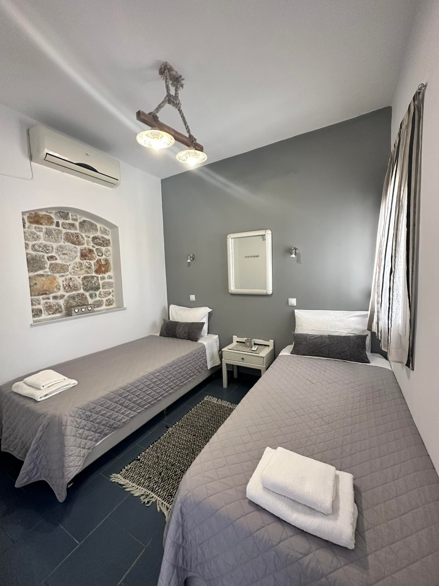 A room with two single beds, a stone wall, a small bedside table with a lamp, and a ceiling light fixture.