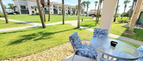 A covered outdoor seating area with a glass table and blue patterned chairs, overlooking a well-maintained lawn with palm trees and a building in the background.