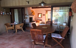 Un comedor con una mesa de madera redonda y seis sillas, una pequeña cocina con estufa y encimera, y un patio exterior accesible por una puerta corrediza de vidrio.