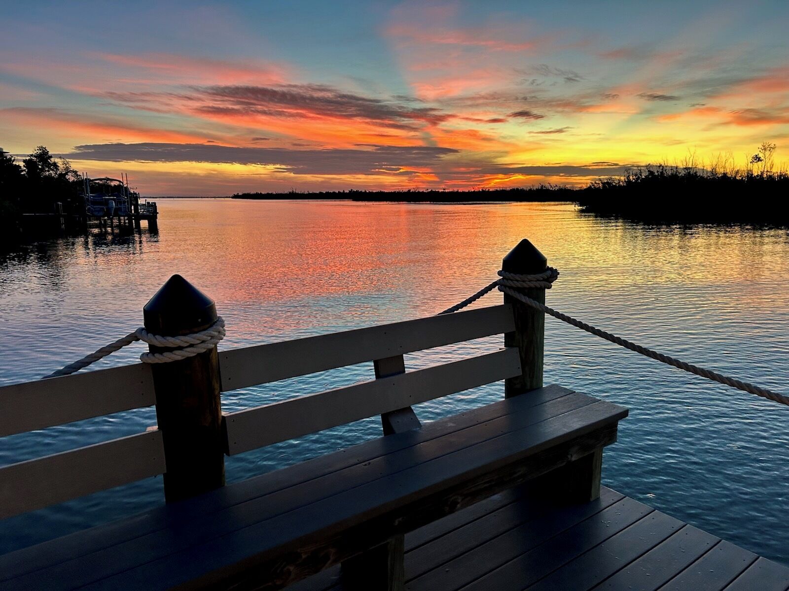 A wooden dock with a railing, ropes, and a clear view of the water and distant shoreline during sunset.
