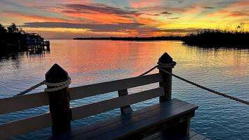 A wooden dock with a railing, ropes, and a clear view of the water and distant shoreline during sunset.