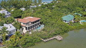 A coastal property with a large house featuring a balcony and a pool, surrounded by lush greenery and a clear view of the ocean. - Bayfront Luxury Home with Private Pool, Spa & Panoramic Views (Captiva)