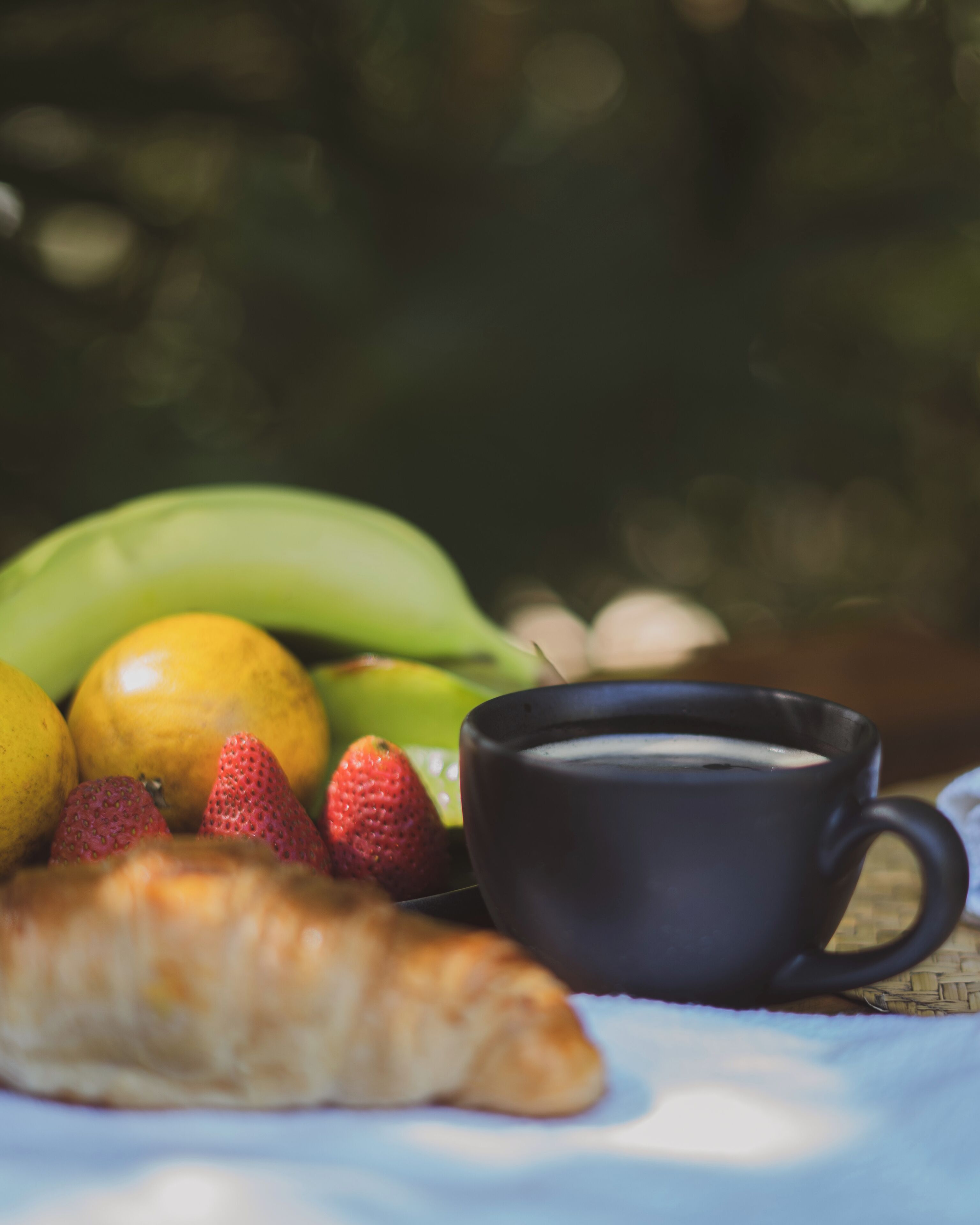 A table with a black cup, a croissant, strawberries, a lemon, and a banana.