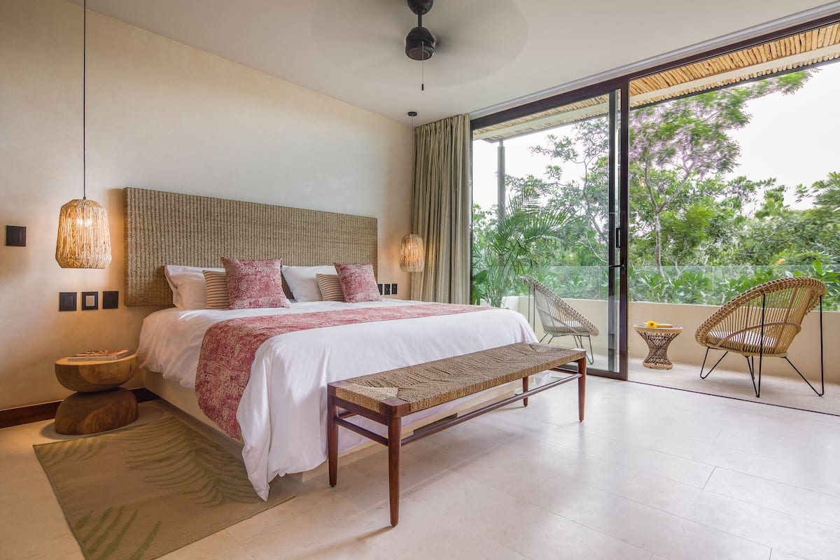 A bedroom with a large bed, a bench, wicker chairs, a small table, and a view of greenery outside.