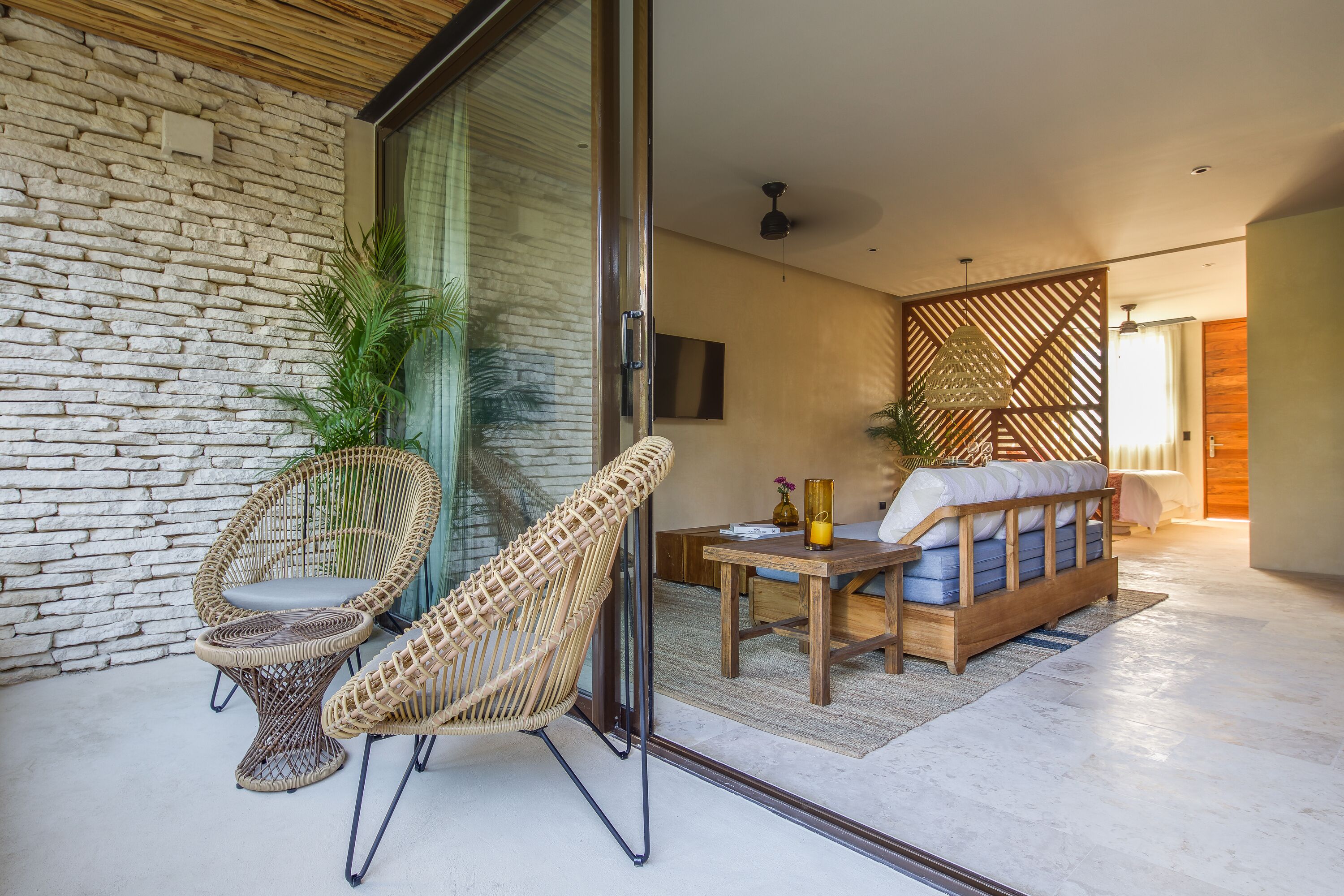 A modern living room with a stone wall, wicker chairs, a wooden sofa, and a glass door leading to a balcony.