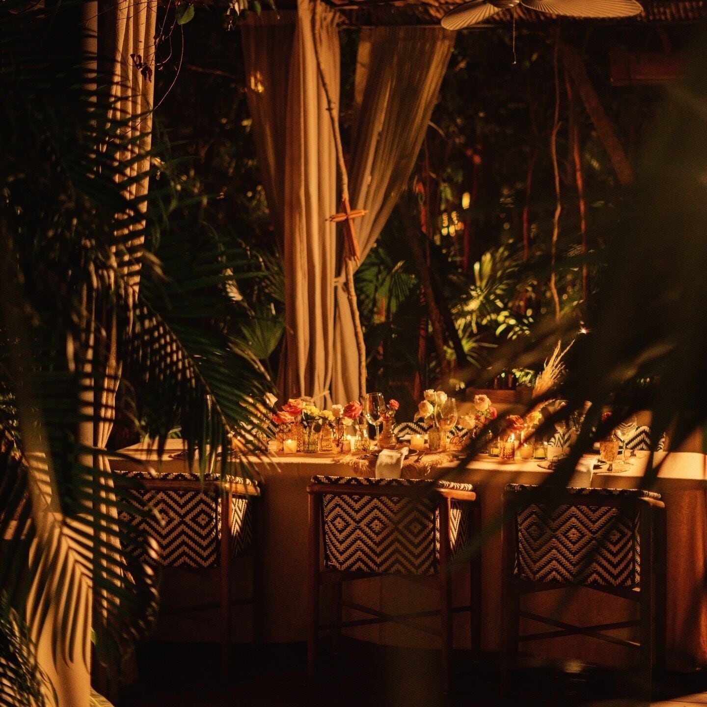 An outdoor dining area with tables set for two, surrounded by lush greenery and draped fabric.