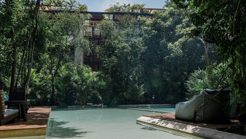 Una piscina rodeada de abundante vegetación, con una terraza de madera y sillones de descanso.