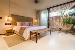 A modern bedroom with a large bed, a bench, wicker chairs, and a view of the outdoors.