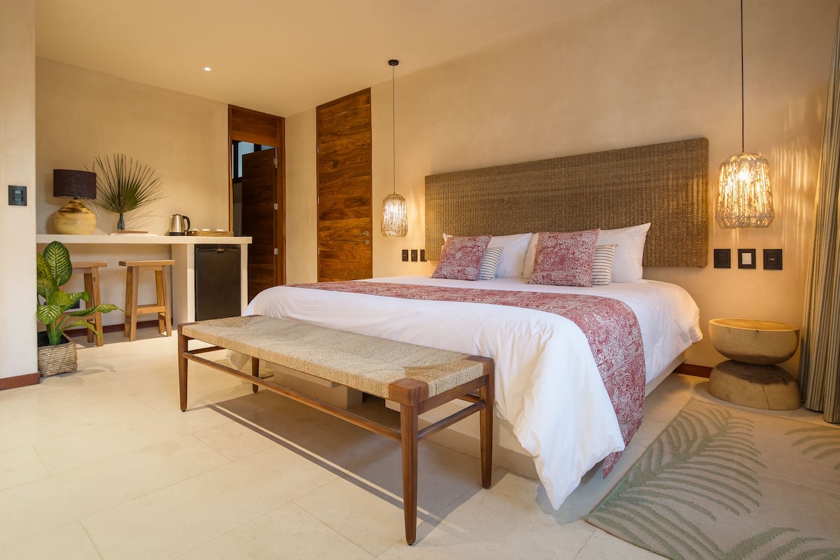 A modern bedroom with a large bed, bedside tables, a bench, and a desk with a lamp and plant.