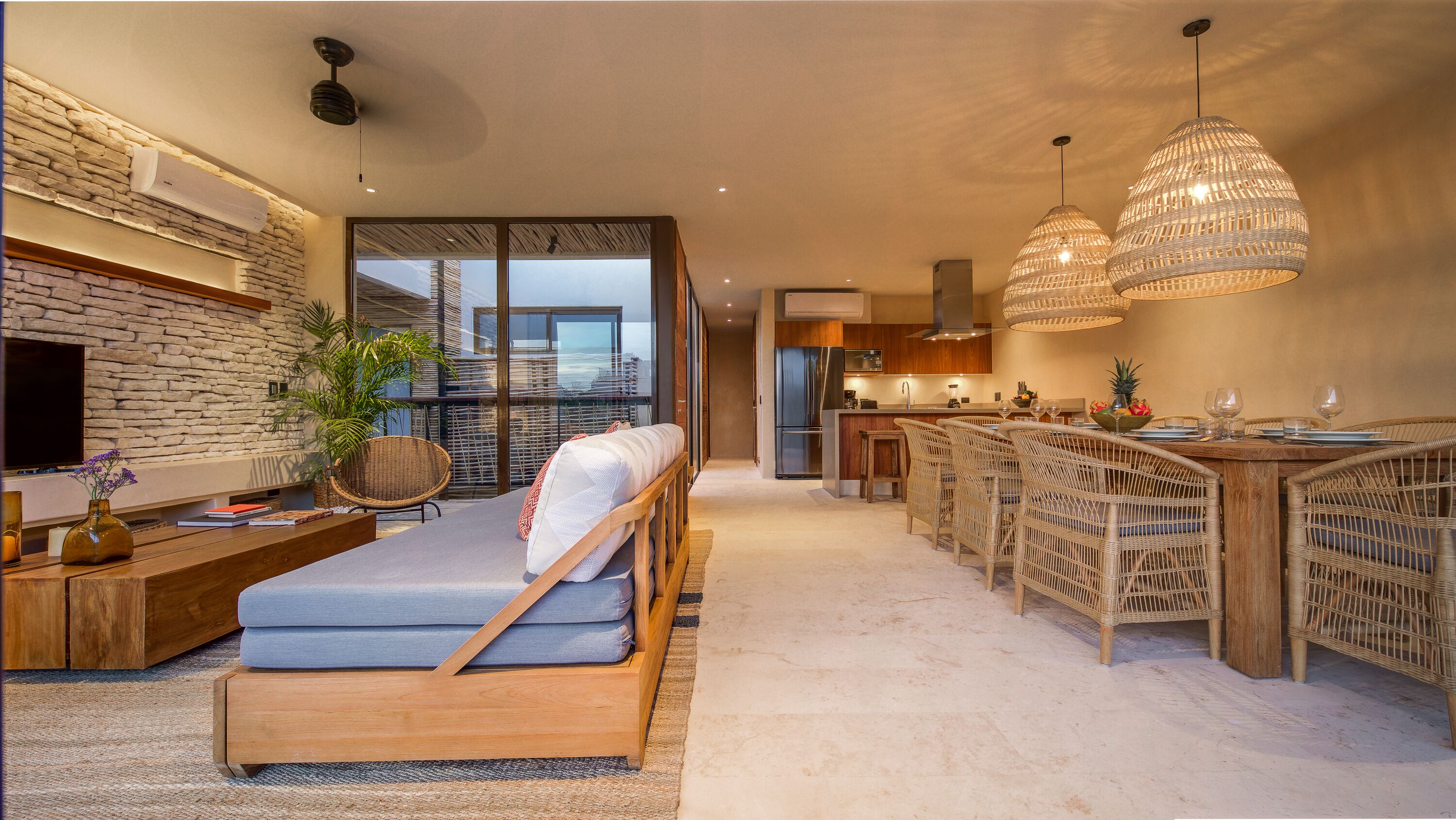 A modern living room with a sofa, a dining area with wicker chairs, a kitchen with a refrigerator, and a large window overlooking a cityscape.