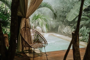 Una terraza de madera con una silla de mimbre, una pequeña piscina y abundante follaje tropical.